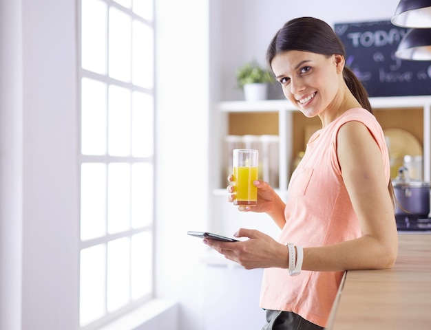 Smiling pretty woman looking at mobile phone and holding glass of orange juice while having breakfas