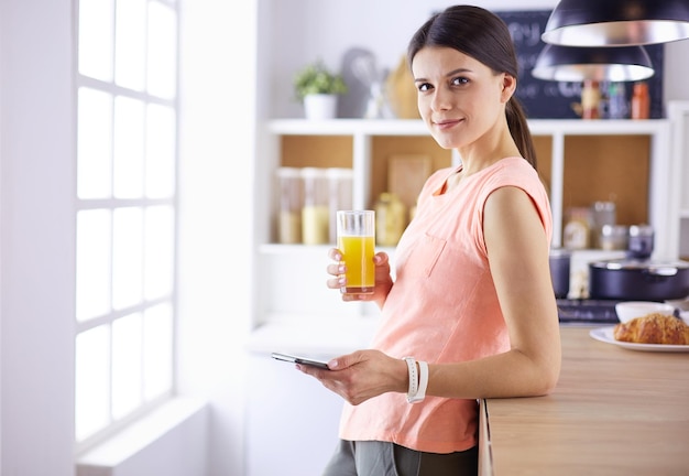 Smiling pretty woman looking at mobile phone and holding glass of orange juice while having breakfast in a kitchen