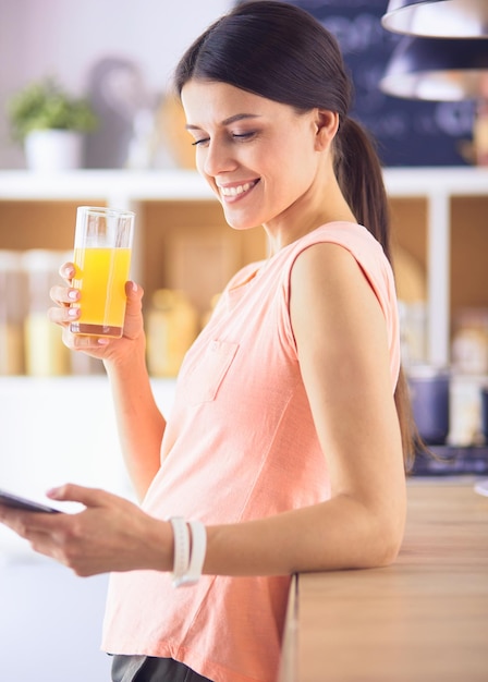 Smiling pretty woman looking at mobile phone and holding glass of orange juice while having breakfast in a kitchen