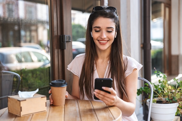 Smiling pretty woman sitting at the cafe outdoors, drinking coffee, using mobile phone