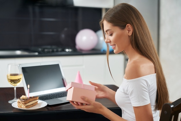 Smiling pretty woman unpacking birthday gift while sitting at home with wireless laptop