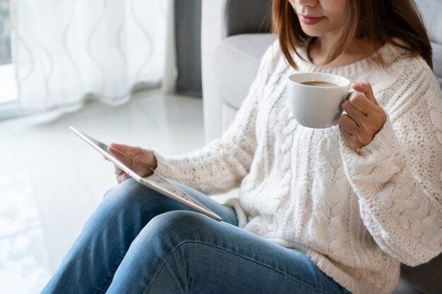 Smiling pretty woman in white sweater using tablet computer while holding a cup of coffee sitting in the living room at home