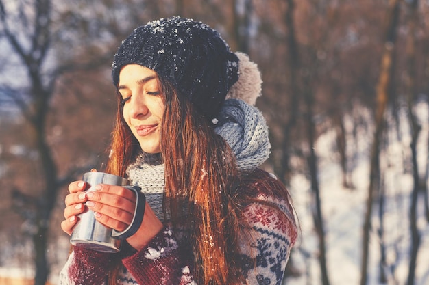 Smiling pretty woman with cup of hot drink standing near winter forest