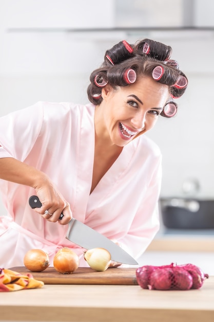 Smiling pretty woman with hair curlers cuts yellow onion on a chopping board in the kitchen.