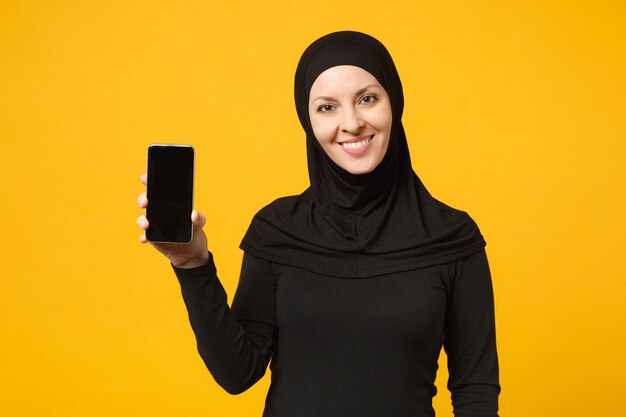 La donna musulmana araba abbastanza giovane sorridente in vestiti neri di hijab tiene in mano il telefono cellulare isolato sul ritratto giallo della parete. concetto di stile di vita religioso della gente. .