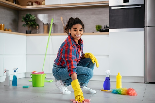 Smiling pretty young black female washes floor with cleaning supplies in modern kitchen interior