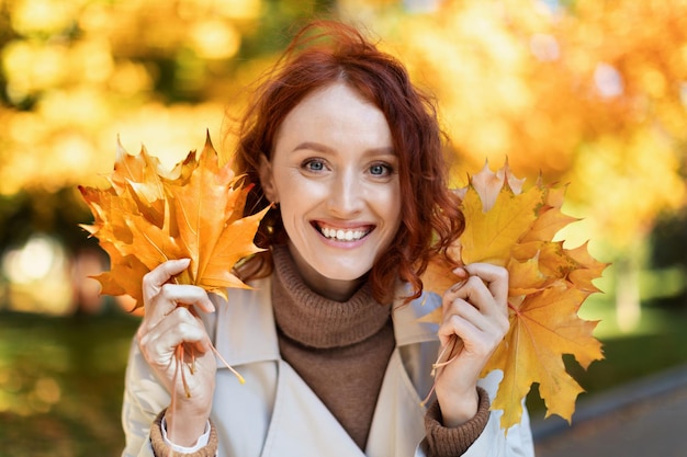 Foto la bella giovane signora europea dai capelli rossi sorridente in impermeabile tiene le foglie gialle si diverte da sola nel parco cittadino