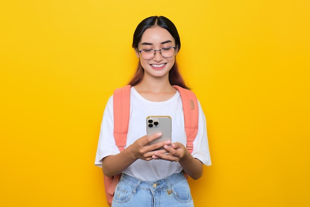 Smiling pretty young student girl wearing backpack and eyeglasses using mobile phone isolated on yellow background