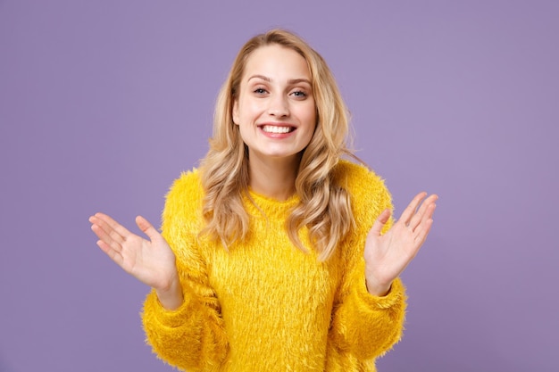 パステル紫の壁の背景に分離された黄色の毛皮のセーターのポーズで笑顔のかなり若い女性の女の子。人々の誠実な感情のライフスタイルの概念。コピースペースをモックアップします。ポインティングハンドを脇に広げます。