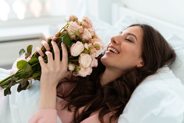 a smiling pretty young woman indoors at home lies in bedroom in bed holding flowers.