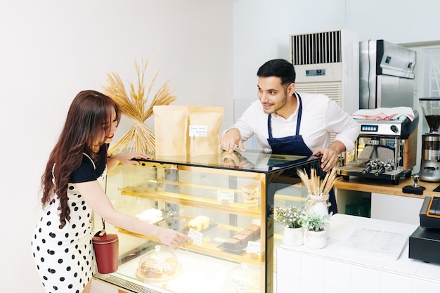 Smiling pretty young woman pointing at showcase and asking waiter to give her a piece of cake