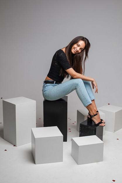 Smiling pretty young woman posing in  while sitting on black boxes, isolated on grey wall