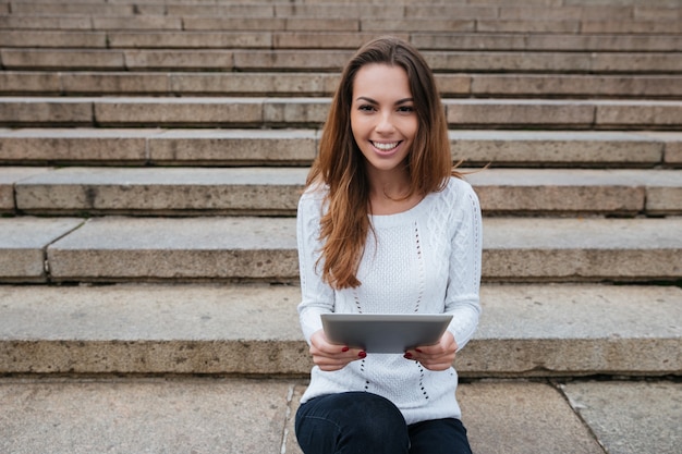 Smiling pretty young woman sitting and using tablet on ladder