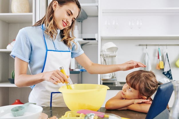 Smiling pretty young woman stroking head of daughter when whisking eggs in big plastic bowl