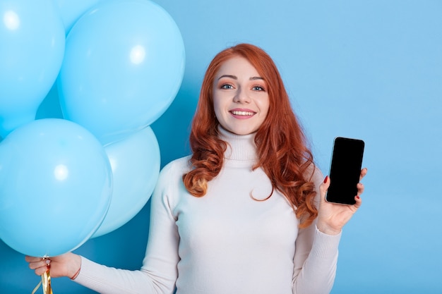 Smiling pretty young woman in white shirt, holds blue balloons and mobile phone with blank empty screen