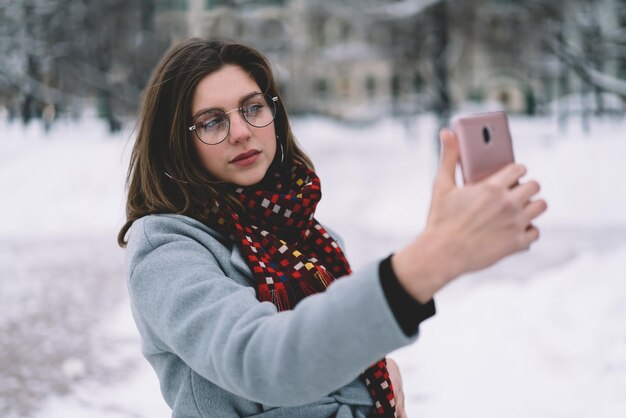 Smiling pretty young woman in winter outfit capturing selfie on\
smartphone at street with snow