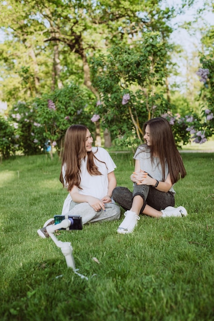 Photo smiling pretty young women influencer bloggers are filming or recording video with their mobile phone on a stabilizer, in a sunny green park outside. blogging concept. soft selective focus.