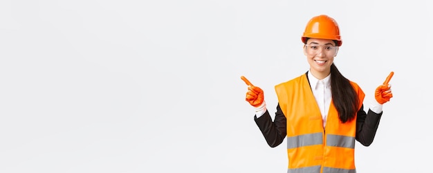 Smiling professional asian female builder engineer in safety helmet protective glasses pointing fingers sideways showing left and right project introduce construction projects white background