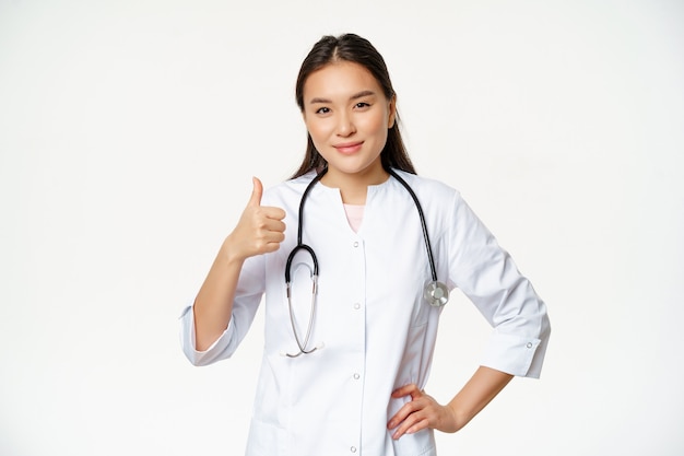 Smiling professional doctor in medical uniform shows thumbs up. Pleased asian female physician confirms smth, recommending product, white background.