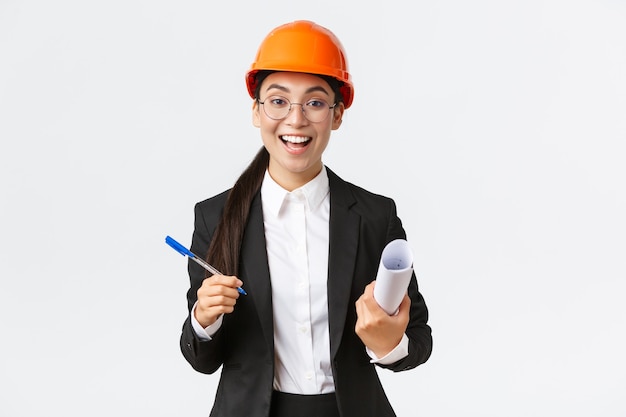 Smiling professional female architect, asian engineer in helmet and business suit, holding blueprints and pen, siging contract, making deal with investors at construction area, white background