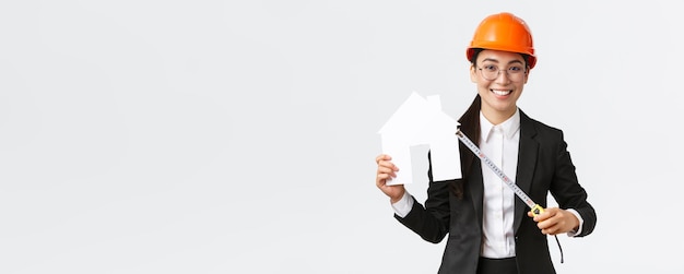 Smiling professional female asian engineer architect in helmet and business suit showing house maket and tape measure ready starting home renovation construction works white background