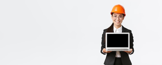 Улыбающаяся профессиональная азиатская женщина-инженер представляет план строительства инвесторам или клиентам, стоящим в защитном шлеме и костюме, показывая экран ноутбука с довольной улыбкой на белом фоне