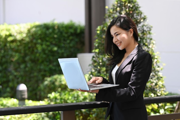 Smiling professional female worker in black suit using laptop while standing outdoor in front of office building