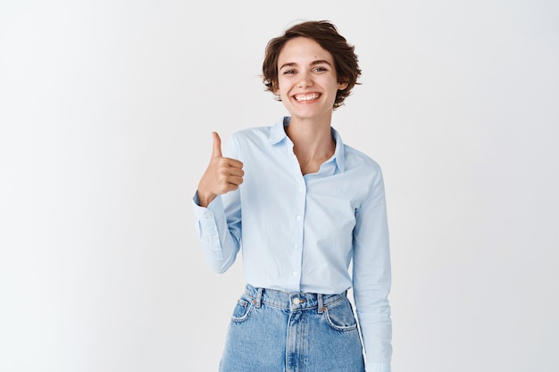 Smiling professional office woman show thumb up, looking satisfied, approve and recommend company, praising good job, say well done, standing on white wall