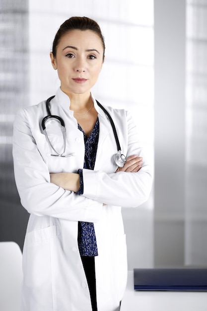 Smiling professional woman-doctor is standing with crossed arms in a clinic office. Portrait of friendly female physician with a stethoscope at work. Perfect medical service in a hospital. Medicine co