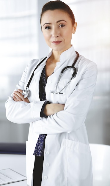 Smiling professional woman-doctor is standing with crossed arms in a sunny clinic office. Portrait of friendly female physician with a stethoscope at work. Perfect medical service in a hospital. Medic