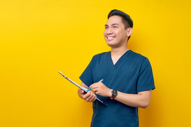 Smiling professional young asian male doctor or nurse wearing a blue uniform holding clipboard and pen writing prescriptions for patients isolated on yellow background healthcare medicine concept