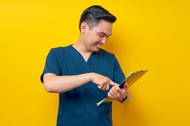 Smiling professional young asian male doctor or nurse wearing a blue uniform pointing finger at clipboard isolated on yellow background healthcare medicine concept