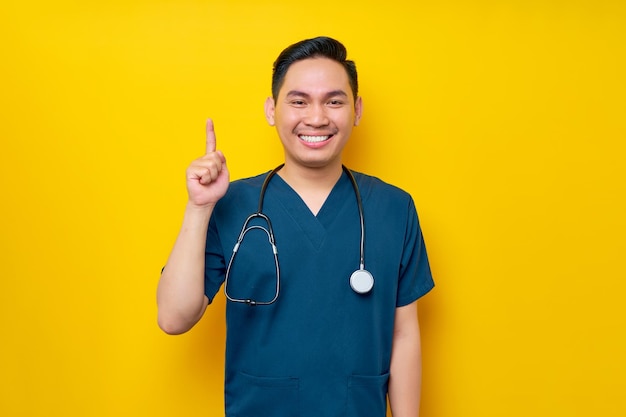 Smiling professional young asian male doctor or nurse wearing a blue uniform pointing finger up isolated on yellow background healthcare medicine concept