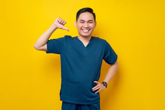 Smiling professional young asian male doctor or nurse wearing a blue uniform standing confident while pointing a finger at himself isolated on yellow background healthcare medicine concept