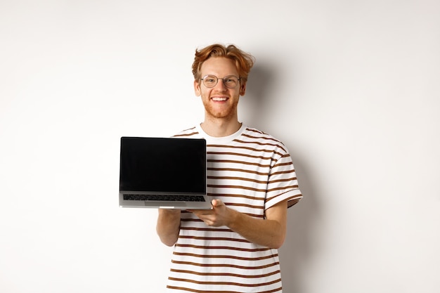 Smiling proud redhead guy showing empty laptop screen. Handsome young man demonstrating logo on computer display, white background.