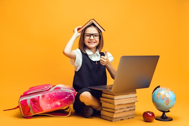 Smiling pupil sits behind a stack of books and pc holds book on head shows thumb up kids education