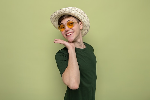 Smiling putting hand under chin young handsome guy wearing hat with glasses isolated on olive green wall