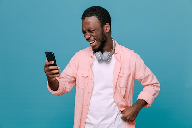 smiling putting hand on hips holding phone young africanamerican guy wearing headphones on neck isolated on blue background