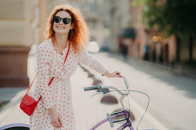 웃고 있는 빨간 머리 유럽 여성은 당신의 승진을 위한 복사 공간이 있는 흐릿한 배경 위에 먼 포즈로 어딘가에 초점을 맞춘 빨간 가방을 들고 자전거 근처에 서 있는 기쁜 표정을 가지고 있습니다
