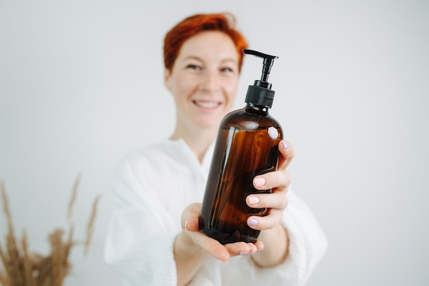 Smiling red haired female chemist presenting a shampoo bottle with a pump. Holding it in front of the camera, focus on the object.