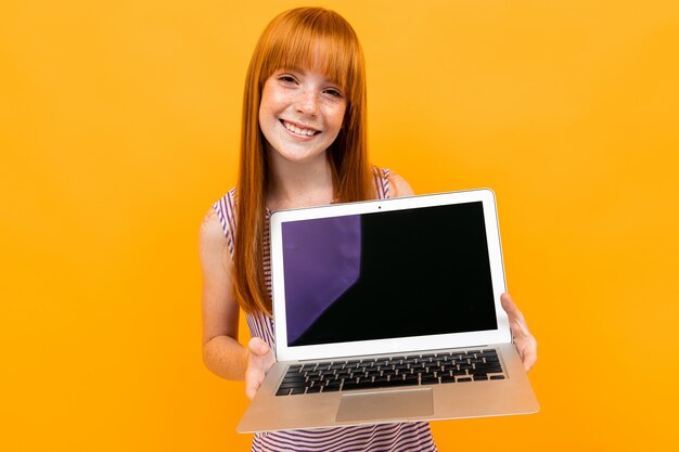 Smiling red-haired girl shows a laptop screen isolated on yellow background