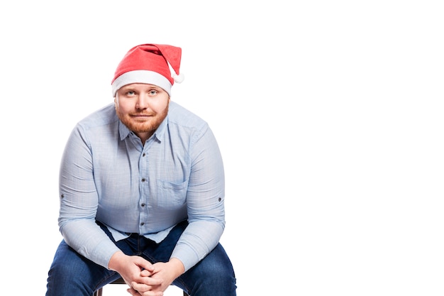 A smiling red-haired man with a beard in a blue shirt, jeans and a Santa Claus hat is sitting on a chair. Close-up. Isolated over white wall. Space for text.