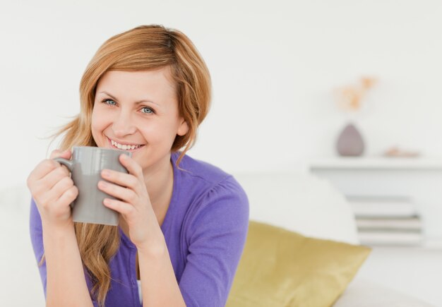 ソファーの上に座っている間、コーヒーを飲んで、ポーズを取っている、笑顔の赤毛の女性