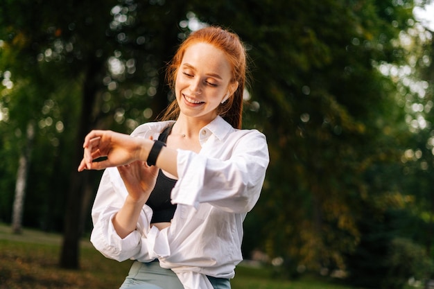 Smiling redhaired young woman is using application on smart watch outdoor in city street at evening