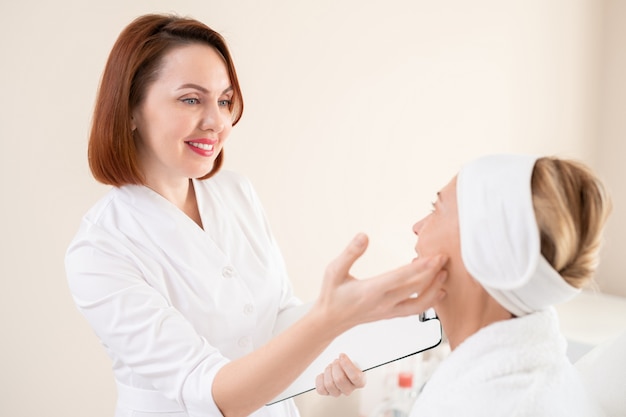 Sorridente cosmetologo rossa con appunti che controlla la pelle del viso del cliente maturo prima della procedura cosmetica