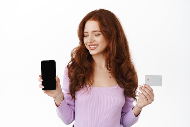 Smiling redhead female model showing credit card, looking at empty smartphone screen, recommending mobile phone app, standing on white