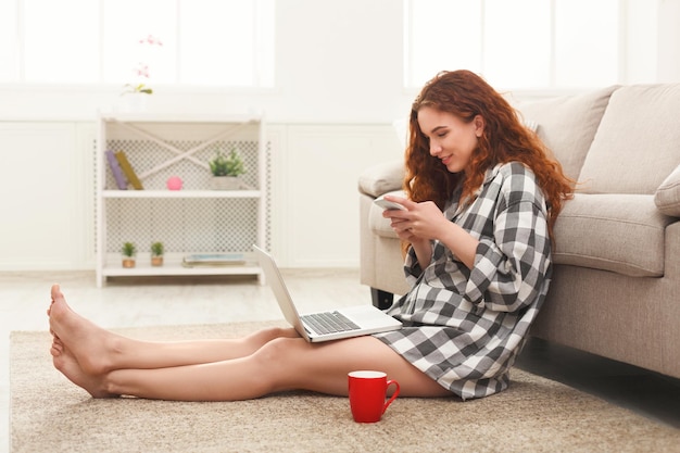 웃는 빨간 머리 소녀는 커피를 마시고 노트북을 사용하고 스마트폰으로 타이핑합니다. 바닥에 앉아 체크 무늬 캐주얼 옷에 곱슬 젊은 여자