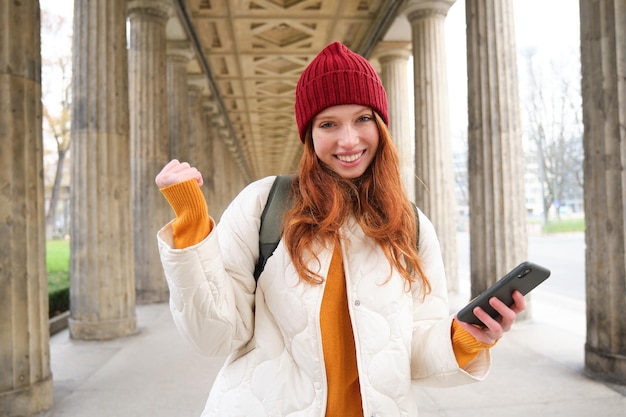Smiling redhead girl looks excited tourist holding mobile phone and does fist pump thrilled about he