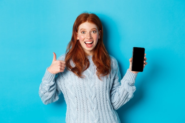 Smiling redhead girl showing smartphone screen, holding phone and demonstrating app, making thumb up in approval, recommending online store, blue background.