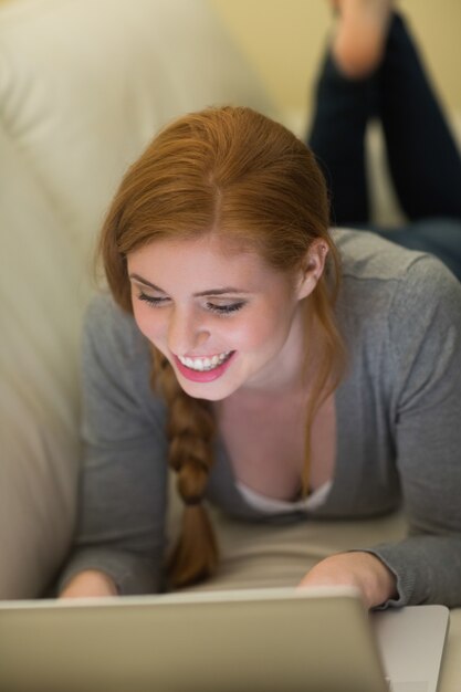 Photo smiling redhead lying on the couch using her laptop at night